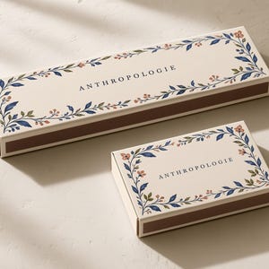 Concept mockup of ivory matchboxes in 28cm and 14.5cm formats, with botanical borders and Anthropologie lettering.