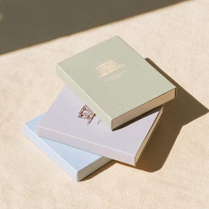 以下が含まれることがあります： 3つの長方形の箱が積み重なっています。一番上の箱はセージグリーン、真ん中はライトラベンダー、一番下はライトブルーです。それぞれの箱の表面には、家の小さな詳細な線画が印刷されています。