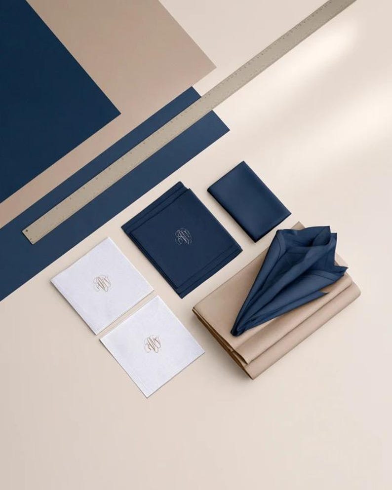 Square navy and cream cocktail napkins with small contrasting monogram letters, displayed with coordinating paper pieces. Digital concept mockup showing a personalised wedding or event table detail.