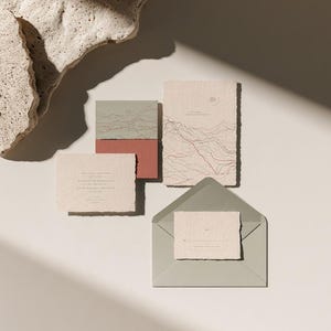 Coordinated wedding invitation stationery with cream cards, muted green accents and an envelope, arranged on a pale background. Fine architectural illustration and small printed text appear on the cards. Digital concept mockup.