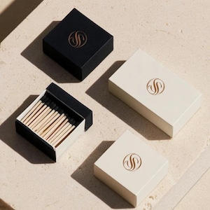 Small rectangular matchbox mockups in a restrained neutral palette, decorated with fine monograms. The styled arrangement shows the paper sleeves and drawer-style construction for personalised event favours.