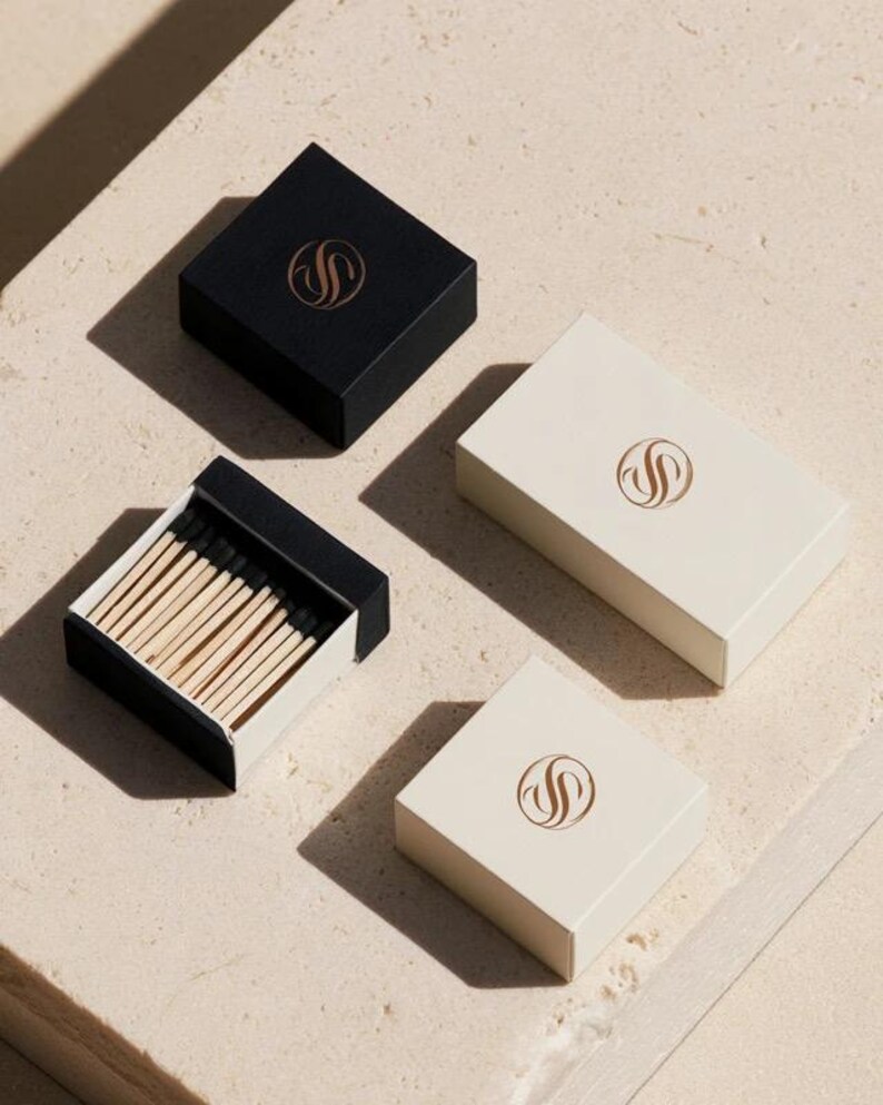 Small rectangular matchbox mockups in a restrained neutral palette, decorated with fine monograms. The styled arrangement shows the paper sleeves and drawer-style construction for personalised event favours.
