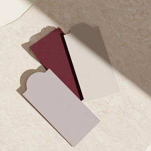 Layered unprinted menu-card mockups with curved paper edges in cream and muted burgundy tones. The arrangement shows contrasting paper shapes and soft shadows for a modern table-stationery design.
