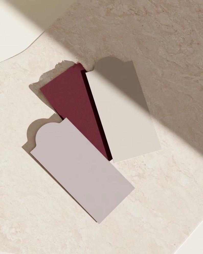 Layered unprinted menu-card mockups with curved paper edges in cream and muted burgundy tones. The arrangement shows contrasting paper shapes and soft shadows for a modern table-stationery design.