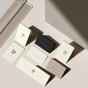 Cream wedding place cards with small dark initials and fine lettering, shown as flat and folded cards on a pale surface. Digital concept mockup of personalised table stationery.