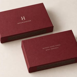 Digital design mockup of two closed burgundy drawer matchboxes in the broad 14.5cm format, showing Houndshire front and reverse artwork in matte ivory standard print on a pale studio surface. No exposed matches; illustrative proportions.
