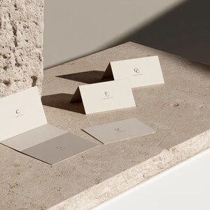 Cream wedding place cards with small dark initials and fine lettering, shown as flat and folded cards on a pale surface. Digital concept mockup of personalised table stationery.