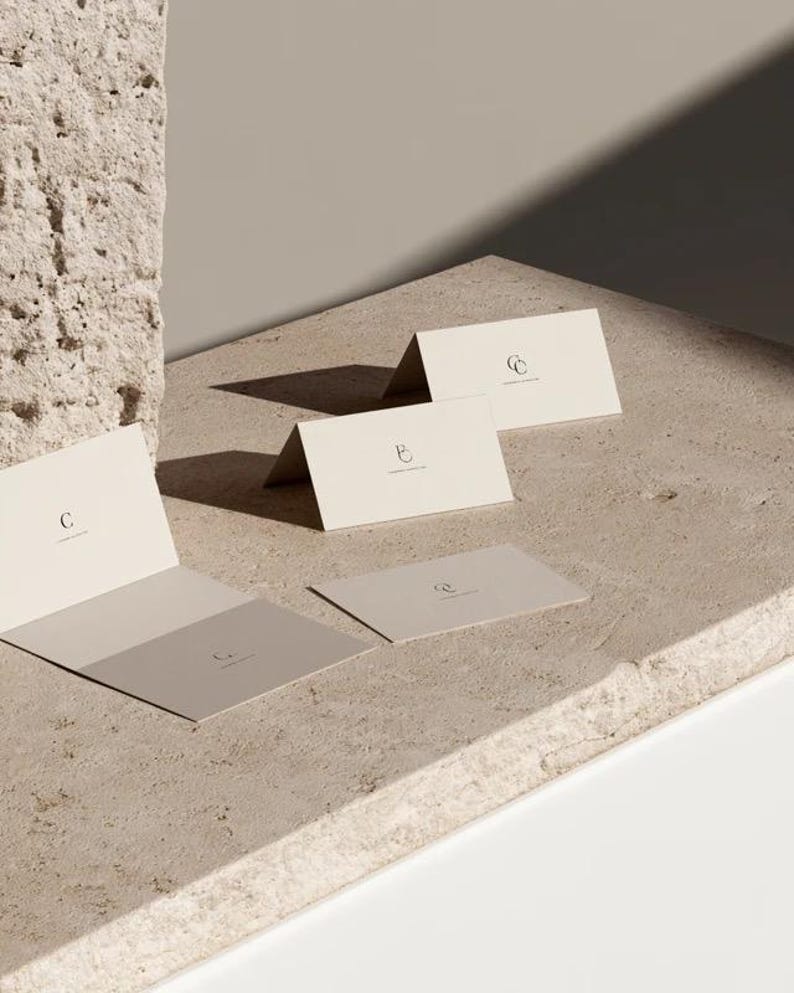 Cream wedding place cards with small dark initials and fine lettering, shown as flat and folded cards on a pale surface. Digital concept mockup of personalised table stationery.