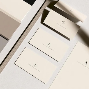 Cream wedding place cards with small dark initials and fine lettering, shown as flat and folded cards on a pale surface. Digital concept mockup of personalised table stationery.