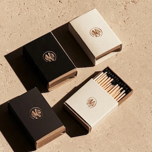 Small rectangular matchbox mockups in a restrained neutral palette, decorated with fine monograms. The styled arrangement shows the paper sleeves and drawer-style construction for personalised event favours.