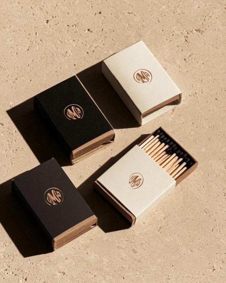 Small rectangular matchbox mockups in a restrained neutral palette, decorated with fine monograms. The styled arrangement shows the paper sleeves and drawer-style construction for personalised event favours.