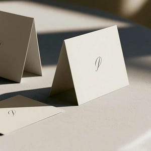 Cream wedding place cards with small dark initials and fine lettering, shown as flat and folded cards on a pale surface. Digital concept mockup of personalised table stationery.