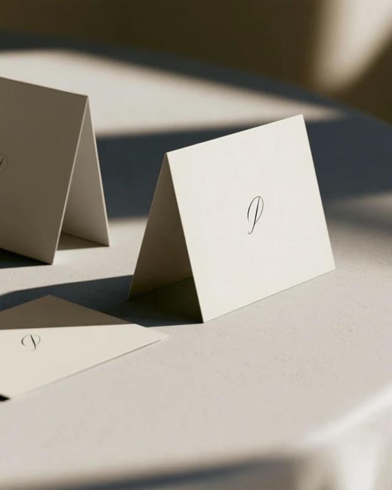 Cream wedding place cards with small dark initials and fine lettering, shown as flat and folded cards on a pale surface. Digital concept mockup of personalised table stationery.