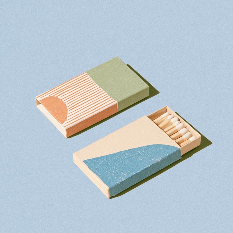 以下が含まれることがあります： 段ボール製の長方形のマッチ箱2つ。1つは閉じており、セージグリーンの天面、ピーチ色の半円、細い白い垂直線が特徴です。もう1つは開いており、白い先端の木製マッチが見えています。
