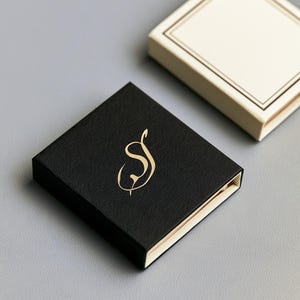 Small rectangular matchbox mockups in a restrained neutral palette, decorated with fine monograms. The styled arrangement shows the paper sleeves and drawer-style construction for personalised event favours.