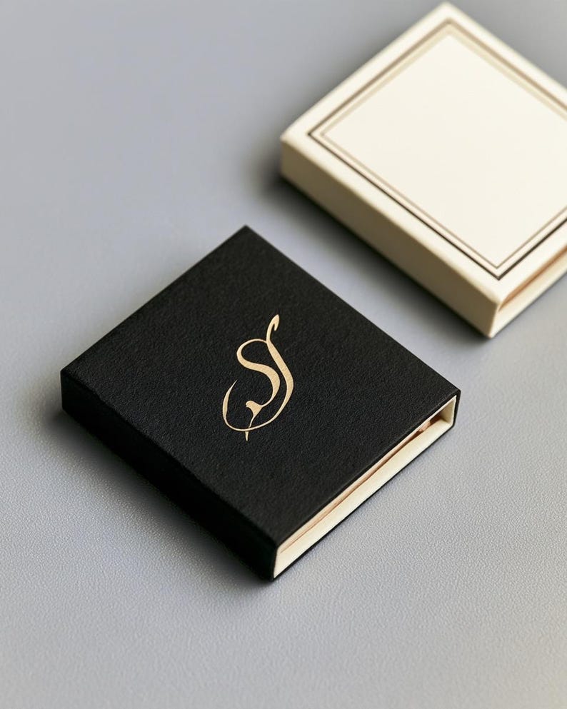 Small rectangular matchbox mockups in a restrained neutral palette, decorated with fine monograms. The styled arrangement shows the paper sleeves and drawer-style construction for personalised event favours.