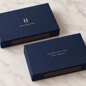 Digital design mockup of closed navy 14.5cm-format drawer matchboxes with gold-foil Houndshire front and reverse branding on pale marble. Foil is an optional paid finish, quoted separately; not a manufactured sample photo.