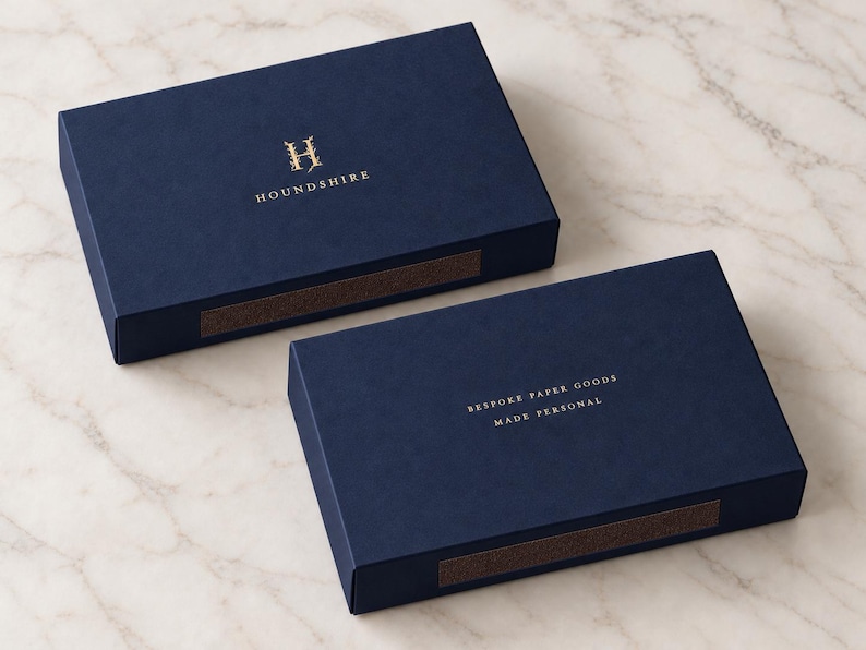 Digital design mockup of closed navy 14.5cm-format drawer matchboxes with gold-foil Houndshire front and reverse branding on pale marble. Foil is an optional paid finish, quoted separately; not a manufactured sample photo.
