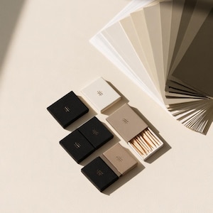 Small rectangular matchbox mockups in a restrained neutral palette, decorated with fine monograms. The styled arrangement shows the paper sleeves and drawer-style construction for personalised event favours.