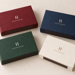 Digital design mockup of the broad 14.5cm matchbox format in burgundy with standard ivory print, navy with gold foil, forest green with silver foil, and ivory with copper foil. All boxes closed; foil examples require a separate paid quote.