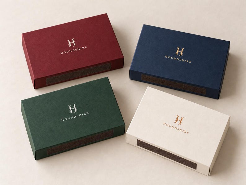 Digital design mockup of the broad 14.5cm matchbox format in burgundy with standard ivory print, navy with gold foil, forest green with silver foil, and ivory with copper foil. All boxes closed; foil examples require a separate paid quote.
