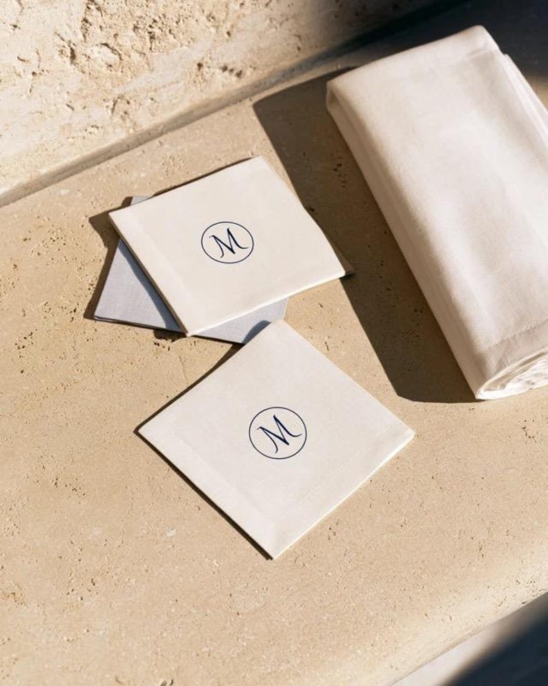 Square navy and cream cocktail napkins with small contrasting monogram letters, displayed with coordinating paper pieces. Digital concept mockup showing a personalised wedding or event table detail.