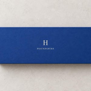 AI-assisted studio illustration of the closed 28 x 10.5 x 3 cm blue matchbox, shown from above with an ivory Houndshire sample monogram. The 28 cm measurement is the outside box length.