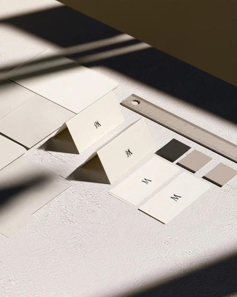 Cream wedding place cards with small dark initials and fine lettering, shown as flat and folded cards on a pale surface. Digital concept mockup of personalised table stationery.