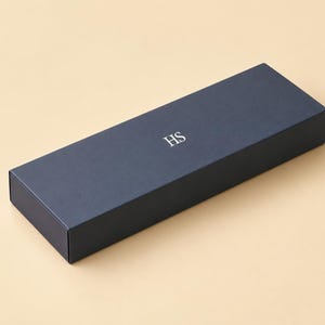 AI-assisted studio design visual of a closed navy sliding matchbox with an example HS monogram, on an ivory background. Illustrates the 28 x 10.5 x 3 cm outside box format; the monogram is an example, not a supplied design.