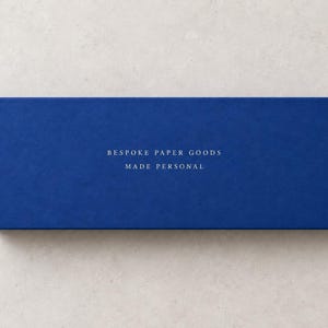 AI-assisted studio illustration of the reverse of the blue 28 cm matchbox, with ivory sample wording: Bespoke Paper Goods, Made Personal.