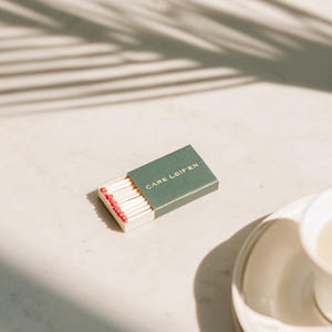 以下が含まれることがあります： 金色の文字で「CARE LOIP'EN」と書かれたダークグリーンのカバーが付いた長方形のマッチ箱。箱は少し開いており、白い紙製のトレイの中に赤い頭の木製マッチが整然と並んでいるのが見えます。