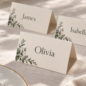 Three folded ivory place cards with green botanical illustrations and the sample names Olivia, James and Isabella, styled on a linen wedding table. Digital concept mockup of personalised guest name cards.