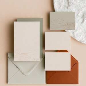 Coordinated wedding invitation stationery with cream cards, muted green accents and an envelope, arranged on a pale background. Fine architectural illustration and small printed text appear on the cards. Digital concept mockup.