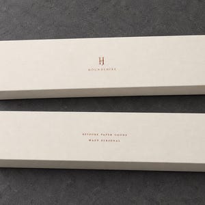 Digital design mockup of two closed ivory extra-long matchboxes with copper-foil Houndshire lettering, showing front and reverse on a charcoal stone surface. Copper foil is an optional upgrade.
