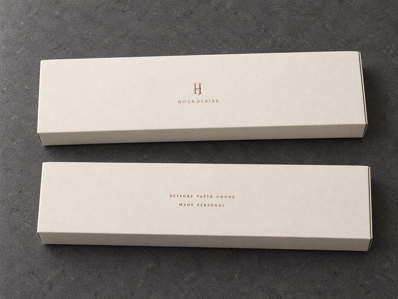Digital design mockup of two closed ivory extra-long matchboxes with copper-foil Houndshire lettering, showing front and reverse on a charcoal stone surface. Copper foil is an optional upgrade.