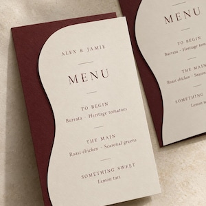 Two finished layered wedding menu cards in ivory and burgundy, with legible sample names Alex & Jamie and a three-course menu in refined serif type. Digital concept mockup showing curved paper layers and printed wording.