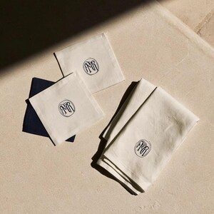 Square navy and cream cocktail napkins with small contrasting monogram letters, displayed with coordinating paper pieces. Digital concept mockup showing a personalised wedding or event table detail.