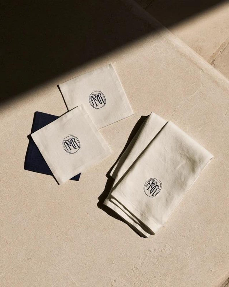 Square navy and cream cocktail napkins with small contrasting monogram letters, displayed with coordinating paper pieces. Digital concept mockup showing a personalised wedding or event table detail.