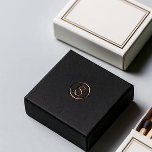 Small rectangular matchbox mockups in a restrained neutral palette, decorated with fine monograms. The styled arrangement shows the paper sleeves and drawer-style construction for personalised event favours.