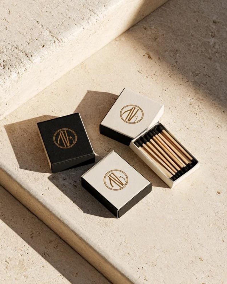 Small rectangular matchbox mockups in a restrained neutral palette, decorated with fine monograms. The styled arrangement shows the paper sleeves and drawer-style construction for personalised event favours.