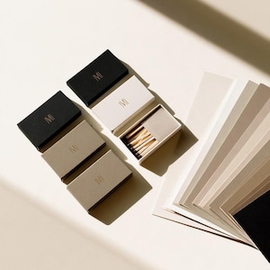 Small rectangular matchbox mockups in a restrained neutral palette, decorated with fine monograms. The styled arrangement shows the paper sleeves and drawer-style construction for personalised event favours.