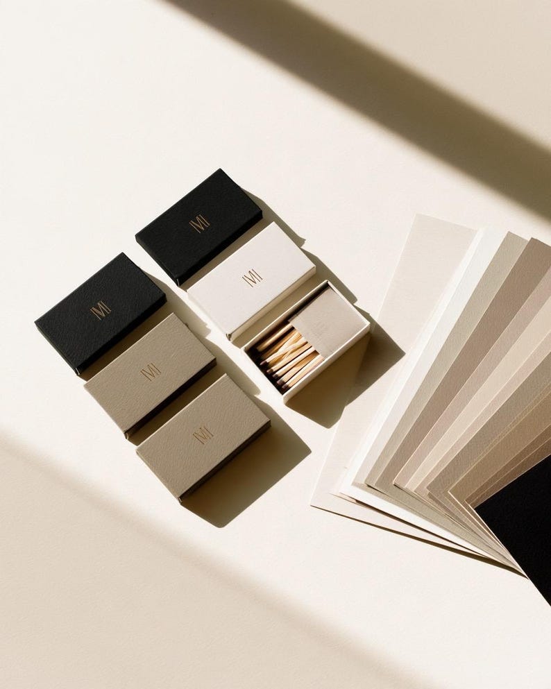 Small rectangular matchbox mockups in a restrained neutral palette, decorated with fine monograms. The styled arrangement shows the paper sleeves and drawer-style construction for personalised event favours.