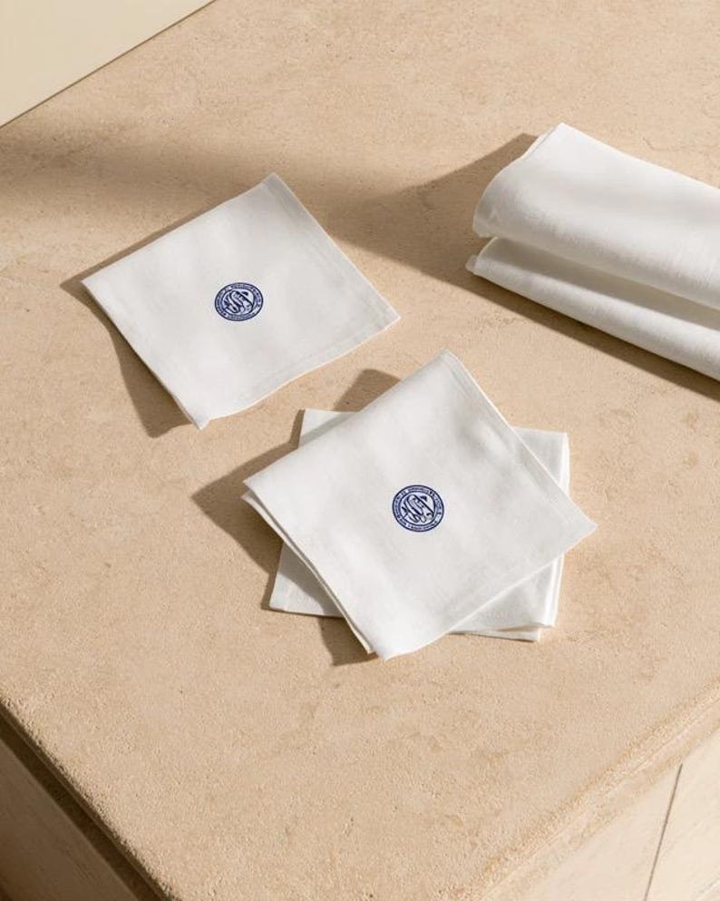 Square navy and cream cocktail napkins with small contrasting monogram letters, displayed with coordinating paper pieces. Digital concept mockup showing a personalised wedding or event table detail.