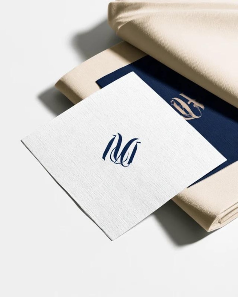 Square navy and cream cocktail napkins with small contrasting monogram letters, displayed with coordinating paper pieces. Digital concept mockup showing a personalised wedding or event table detail.