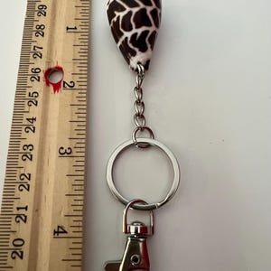 May include: A keychain featuring a brown and white patterned seashell charm attached to a silver-toned metal ring and lobster clasp. A wooden ruler is positioned alongside the keychain, showing the shell measures approximately 1.5 inches.