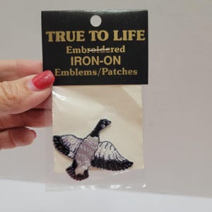 May include: An embroidered iron-on patch of a goose in flight with black, white, and gray thread details. The patch is sealed in a clear plastic bag with a black header card featuring the gold text TRUE TO LIFE Embroidered IRON-ON Emblems/Patches.
