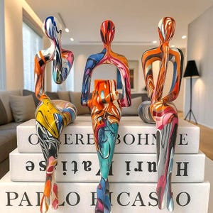 Pode incluir: Três esculturas abstratas em forma humana com um acabamento de pintura marmorizada multicolorida e vibrante. Cada figura está sentada, apresentando membros alongados e formas fluidas. As esculturas estão dispostas sobre uma pilha de três livros brancos.