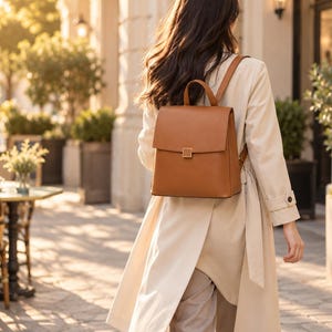 May include: A tan leather backpack with a structured rectangular shape, a top handle, and a flap closure featuring a small square gold-tone metal clasp. The backpack is worn with adjustable shoulder straps over a light beige trench coat.