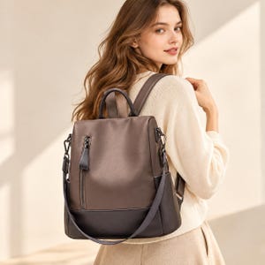 May include: A two-tone brown backpack featuring a smooth, lighter brown upper section and a darker brown base. The bag includes a vertical zippered front pocket with a matching pull tab, a top handle, and adjustable shoulder straps with metal hardware.