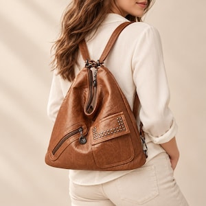 May include: A brown backpack with a textured finish, featuring a top zip closure, a diagonal zipped pocket on the front, and a rectangular flap pocket adorned with small metallic studs. The bag includes thin shoulder straps and dark hardware.